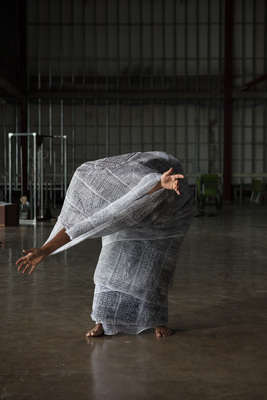 Still of a performance of a person in a newspaper suit so that only their feet and forearms and hands are exposed. They're mid-bow